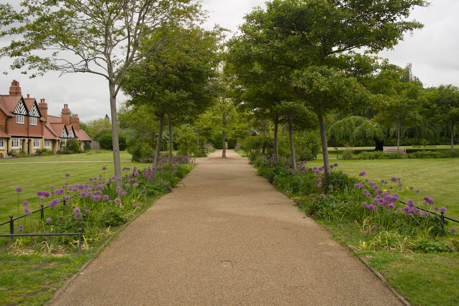 A landscaped town garden featuring a variety of colorful seasonal flower beds, including vibrant red, pink, and white blooms arranged in neat borders around a central stone planter. The garden is bord