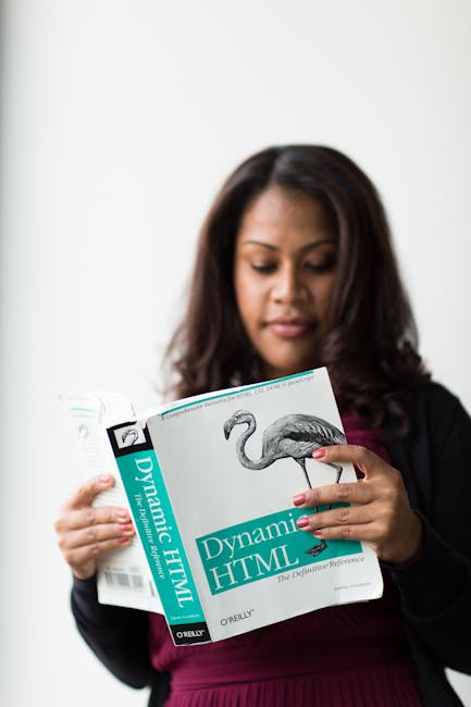 The image shows a woman with dark brown hair and light makeup, wearing a black blazer over a dark pink top, standing indoors against a plain white background. She is holding a book titled 'Dynamic HTM