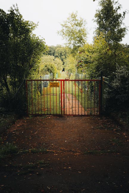 A rusted red metal gate blocking a narrow dirt pathway that leads into a lush, green garden. The pathway is flanked by tall, dense hedges and trees with various shades of green foliage, some with ligh